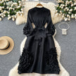 Black dress with ruffled sleeves and bottom on a hanger against a decorative background with flowers and a hat.